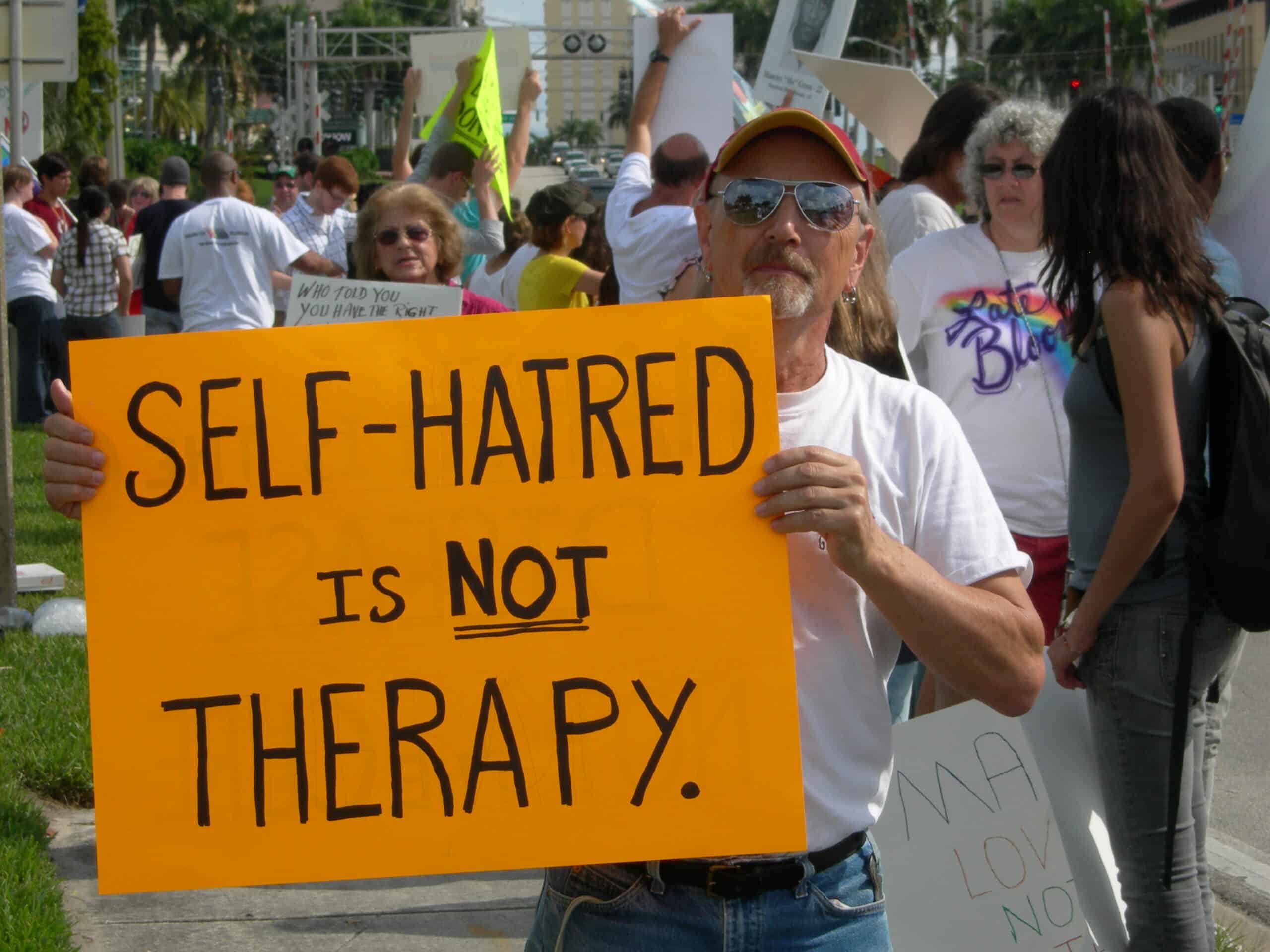 Man protesting with sign that says "Self Hatred is Not Therapy"