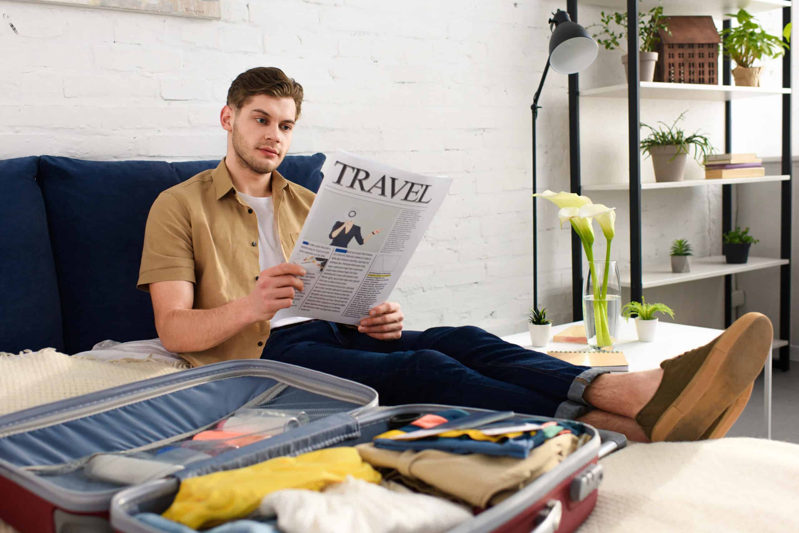 Young man reading travel newspaper while packing suitcase on bed