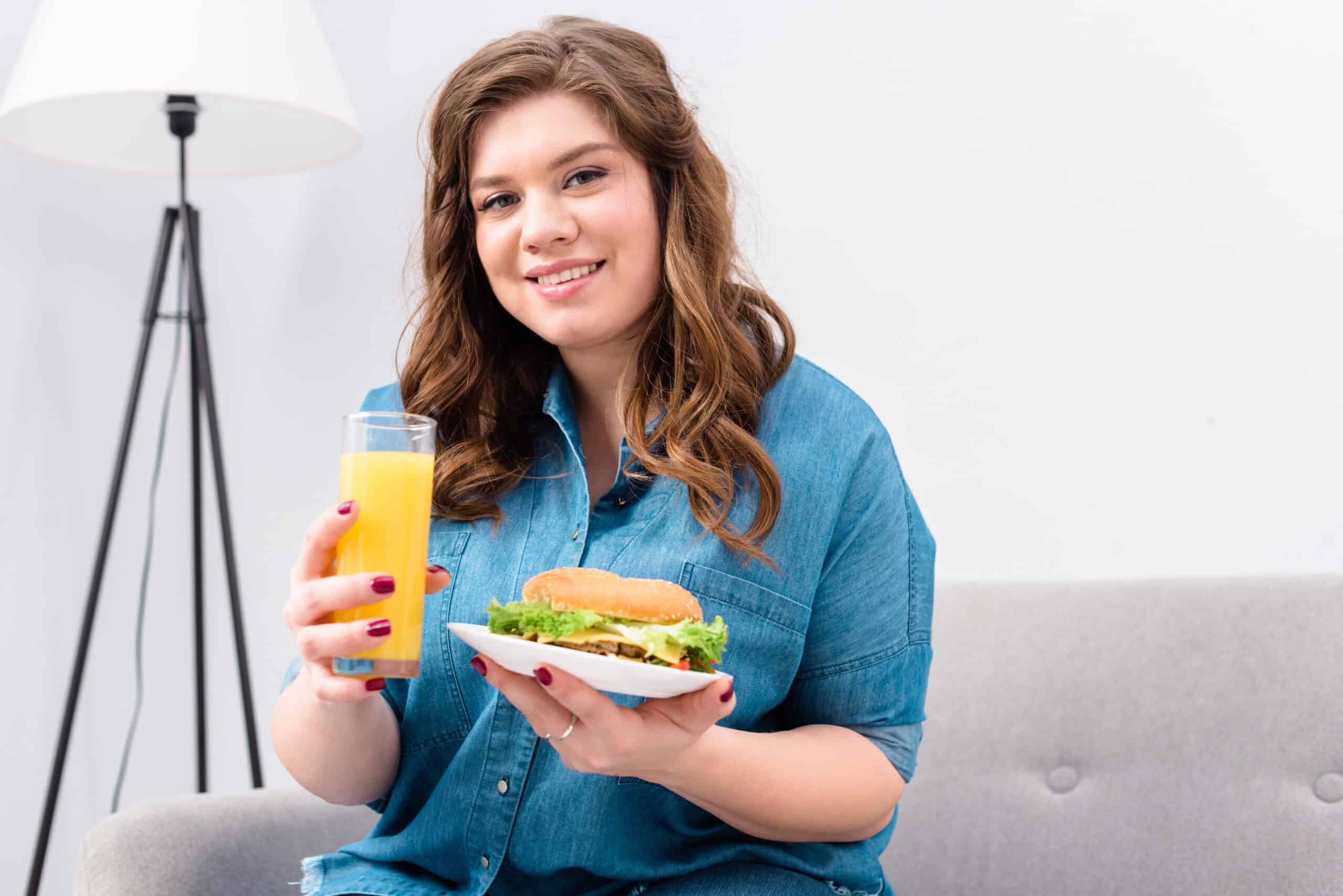 Overweight smiling woman with glass of juice and burger on plate in hands at home