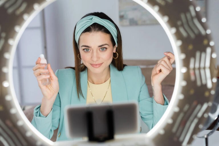 Pretty woman holding spray bottle of micellar water near smartphone in holder with ring light