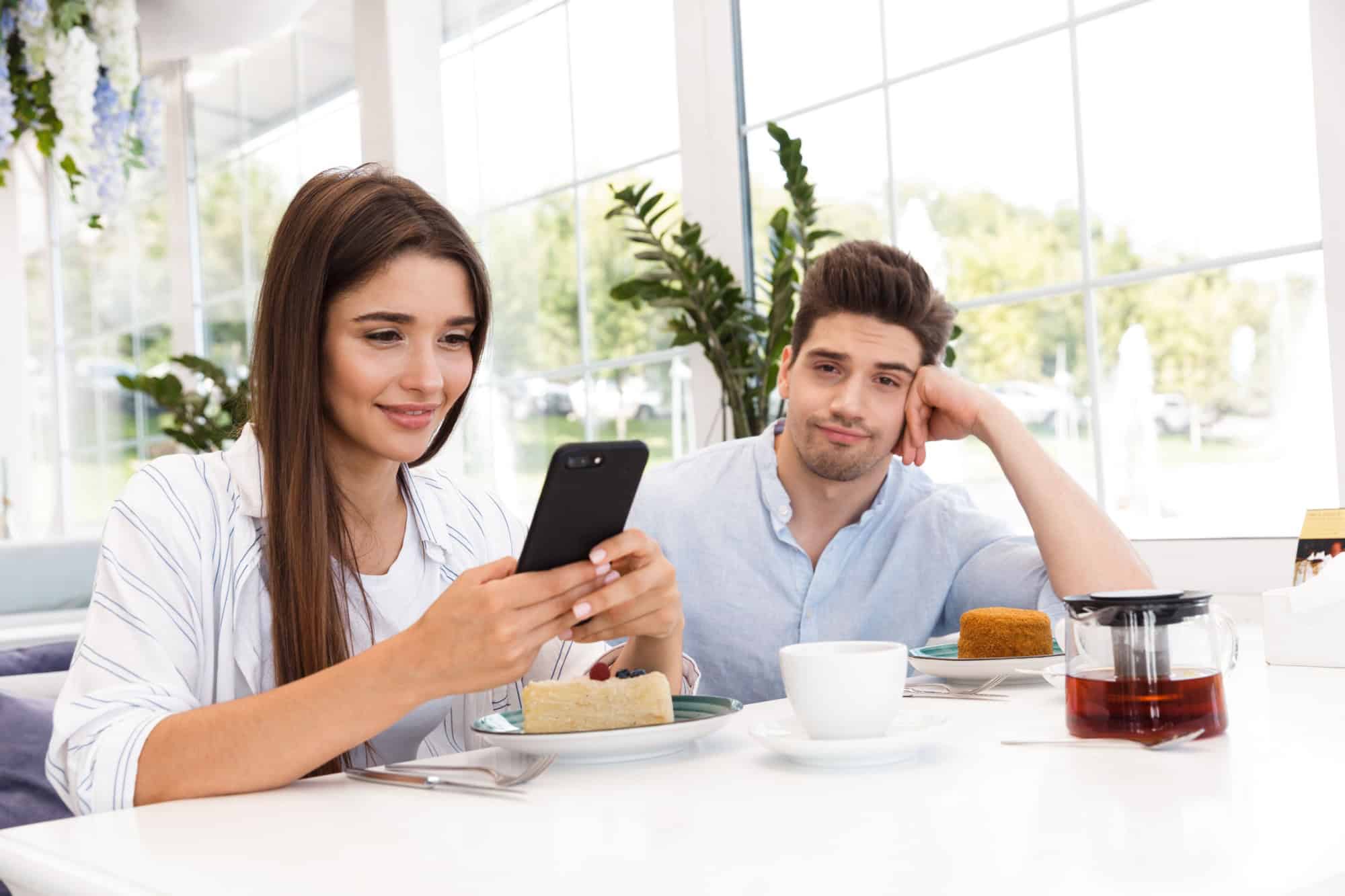 Smiling young man sitting at the cafe table while his girlfriend using mobile phone