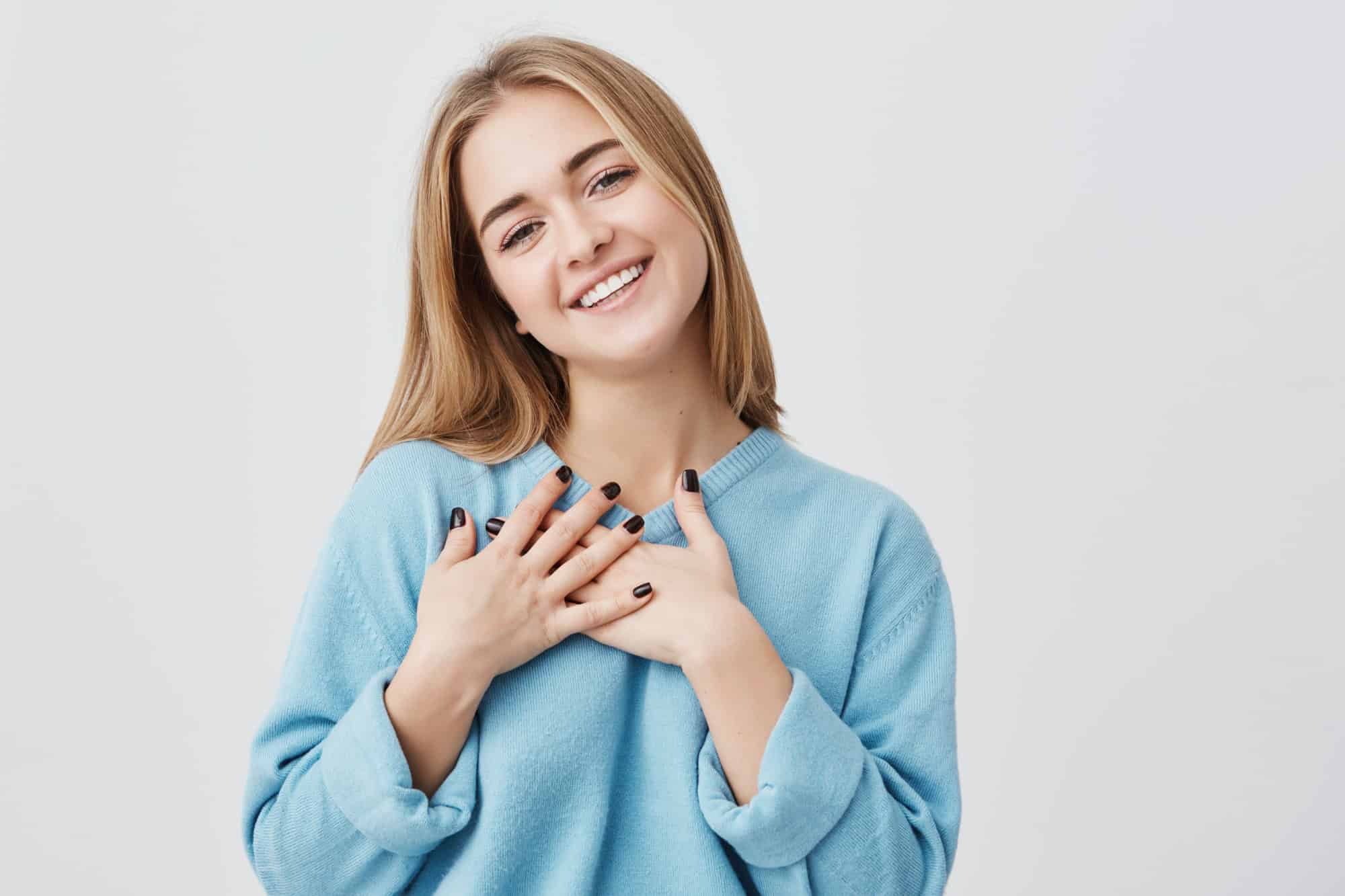 Beautiful positive friendly-looking young european girl with lovely sincere smile feeling thankful and grateful, showing her heart filled with love and gratitude holding hands on her breast