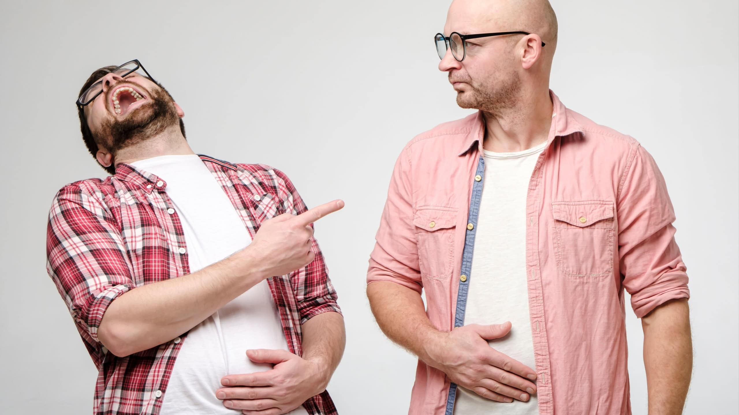 Man laughs out loud, holds on to stomach and points a finger at another person who is upset by the evil joke of his friend.