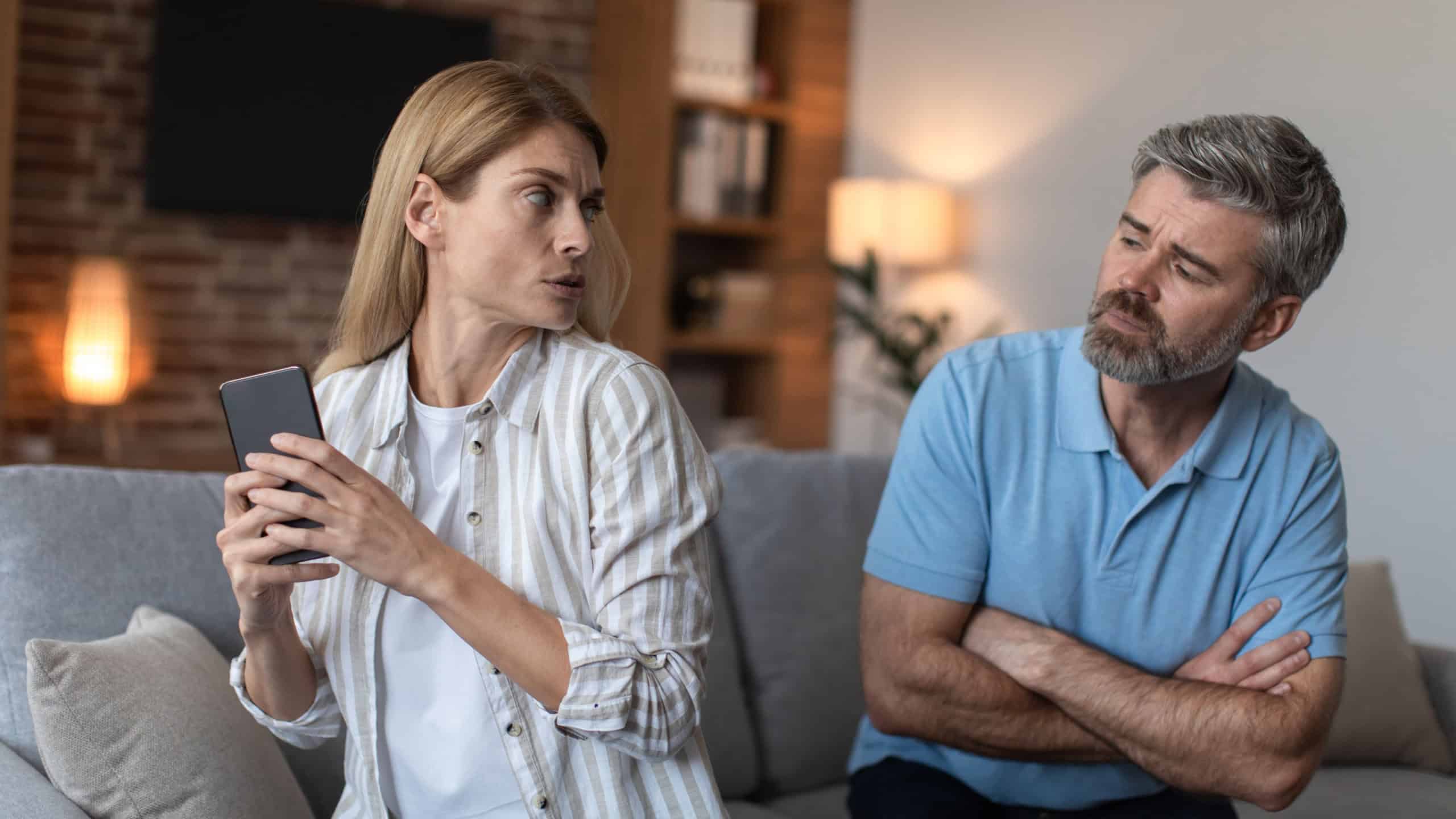 Sad middle aged european man looking at scared woman with smartphone in living room interior.