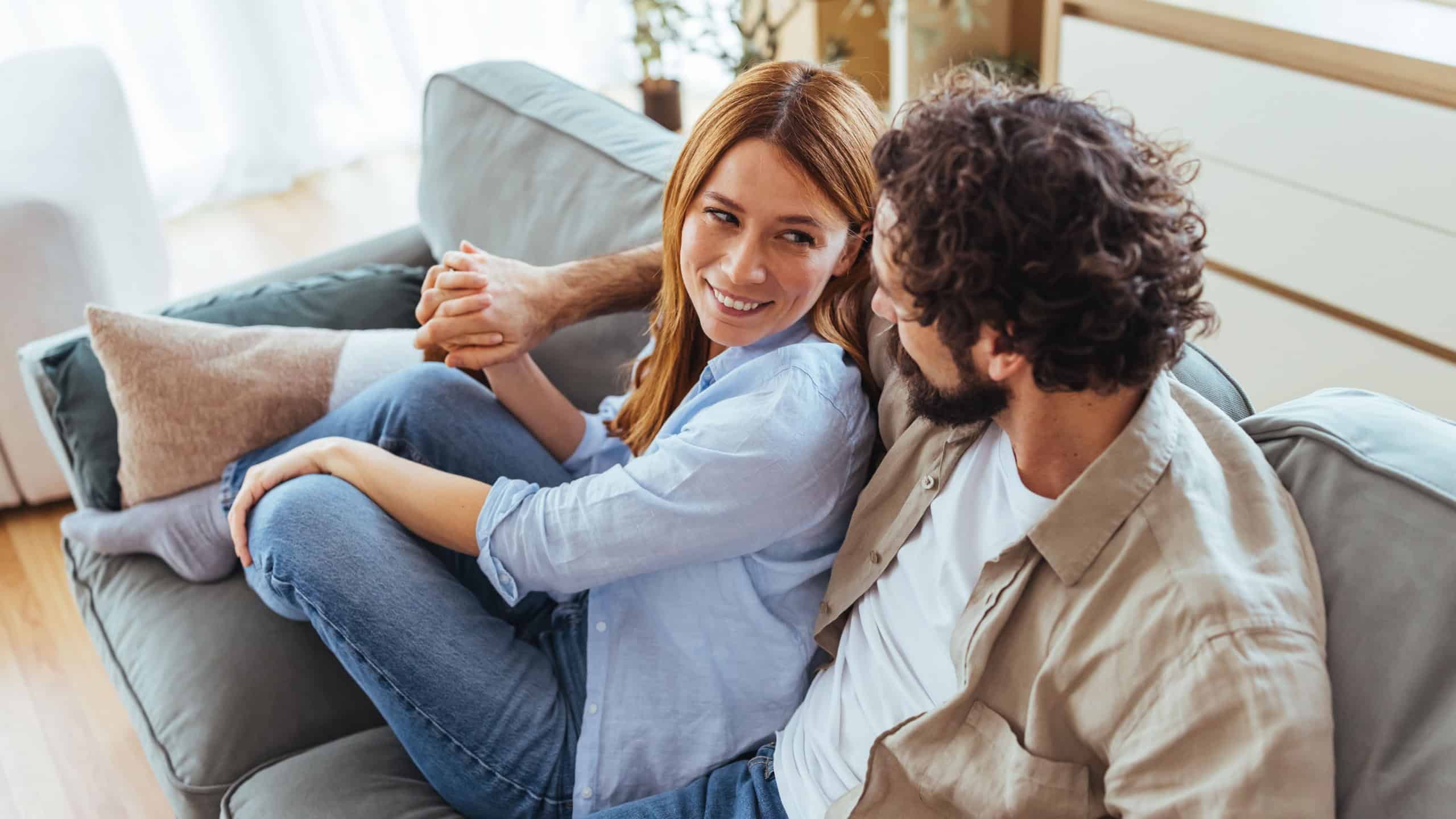 A smiling couple shares a relaxed moment on a sofa in a bright, modern living room. Casual attire and warm light convey comfort, friendship, and everyday togetherness.