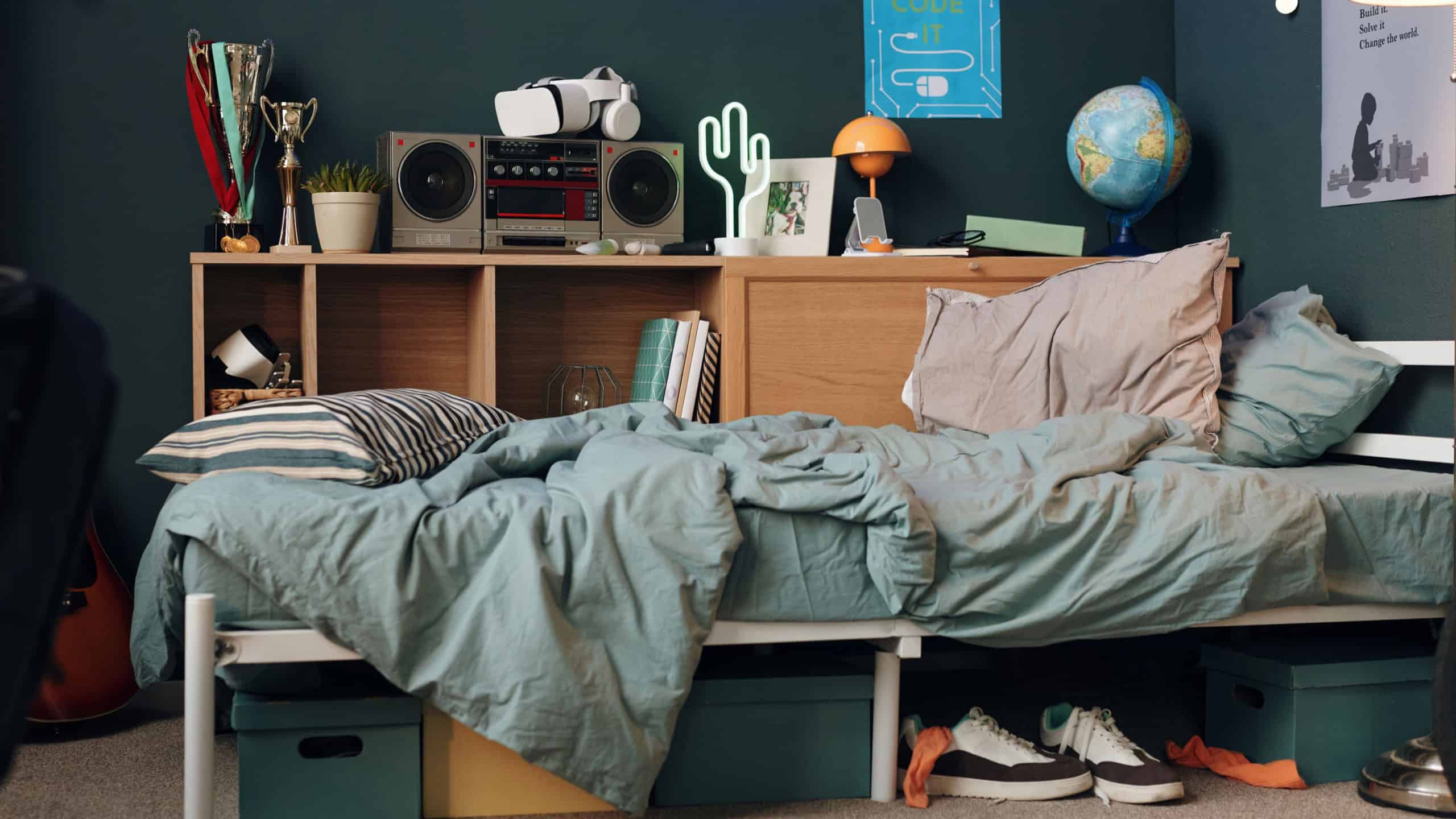 Messy bed with striped pillow and wrinkled blanket in teenager bedroom, bookshelf holding trophies, books, virtual reality headset, radio, globe, sneakers and boxes under bed