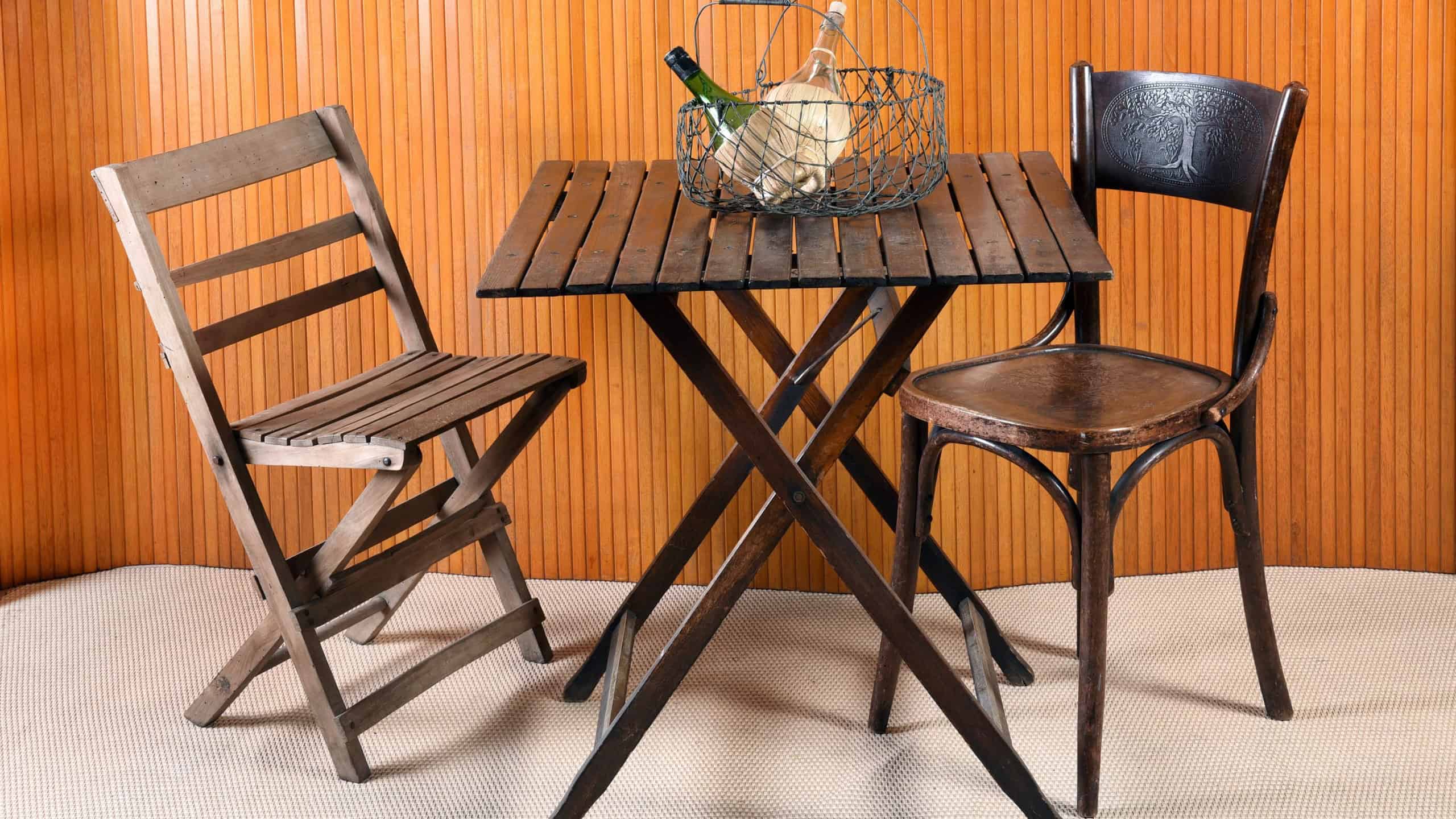 Furniture Still Life of Folding Table with Mismatched Chairs and Wire Basket with Bottles in Room with Beige Carpet and Wood Wall Paneling