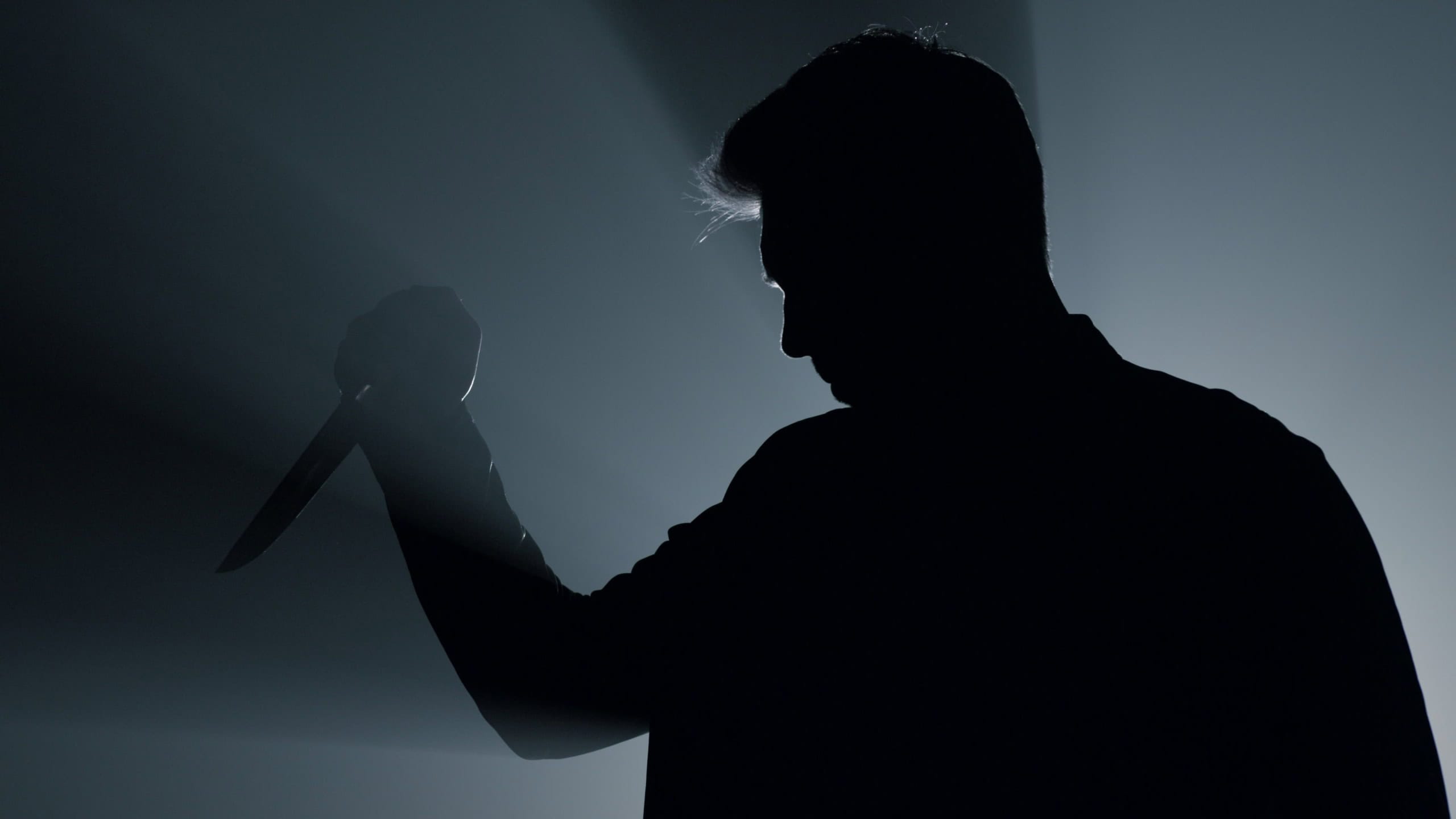 Silhouette of aggressive man standing with knife in hand indoors. Dangerous criminal using weapon in floodlight background. Scary gangster attacking with dagger in dark space. Crime concept.