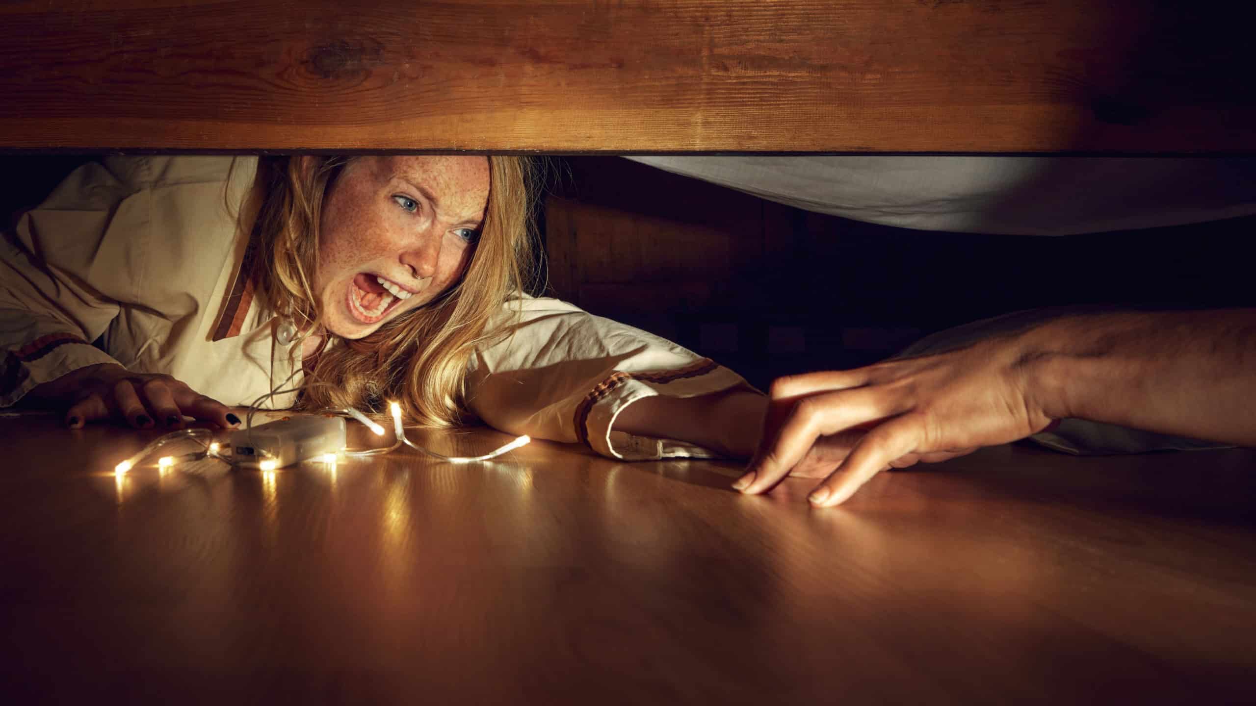 Young fearful woman lying under bed with light while male hand grabbing her. Woman shouting in fear and horror. Spooky season. Concept of Halloween, human emotions