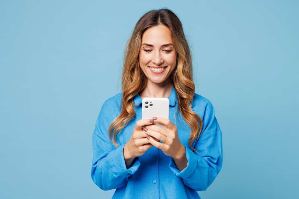 Young smiling happy cheerful woman she wear shirt casual clothes hold in hand use mobile cell phone chat online isolated on plain pastel light blue cyan background studio portrait. Lifestyle concept