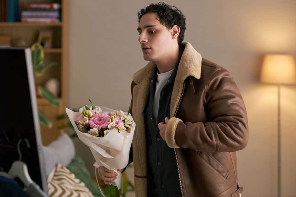 Young adult Caucasian man holding bouquet of flowers preparing for date standing indoors looking at mirror adjusting jacket appearing thoughtful in home setting