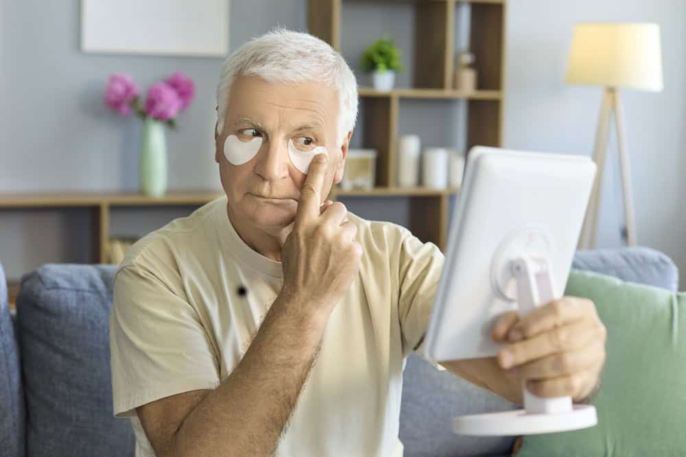 Senior man with patches, using mirror, home skincare. Under eye pads ease puffiness and circles as he perfects a face grooming routine, promoting aging and hygiene. Concept of male eye care.